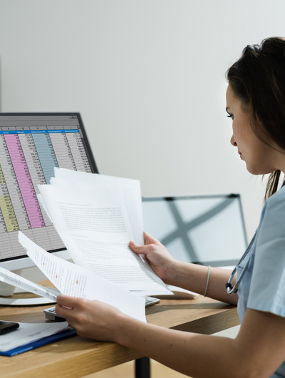 A person in light-blue scrubs with a stethoscope around their neck is seated at a desk, intently reviewing several paper documents. In the background, a computer monitor displays what appears to be a spreadseet or data table.
